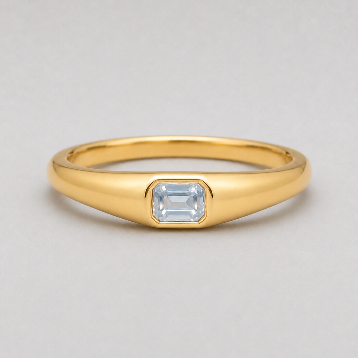 3D render of a polished yellow gold ring featuring a rectangular emerald-cut white sapphire in a bezel setting, shown on a neutral light grey background.