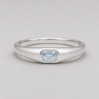 3D render of a polished silver ring featuring a rectangular emerald-cut white sapphire in a bezel setting, shown on a neutral light grey background.