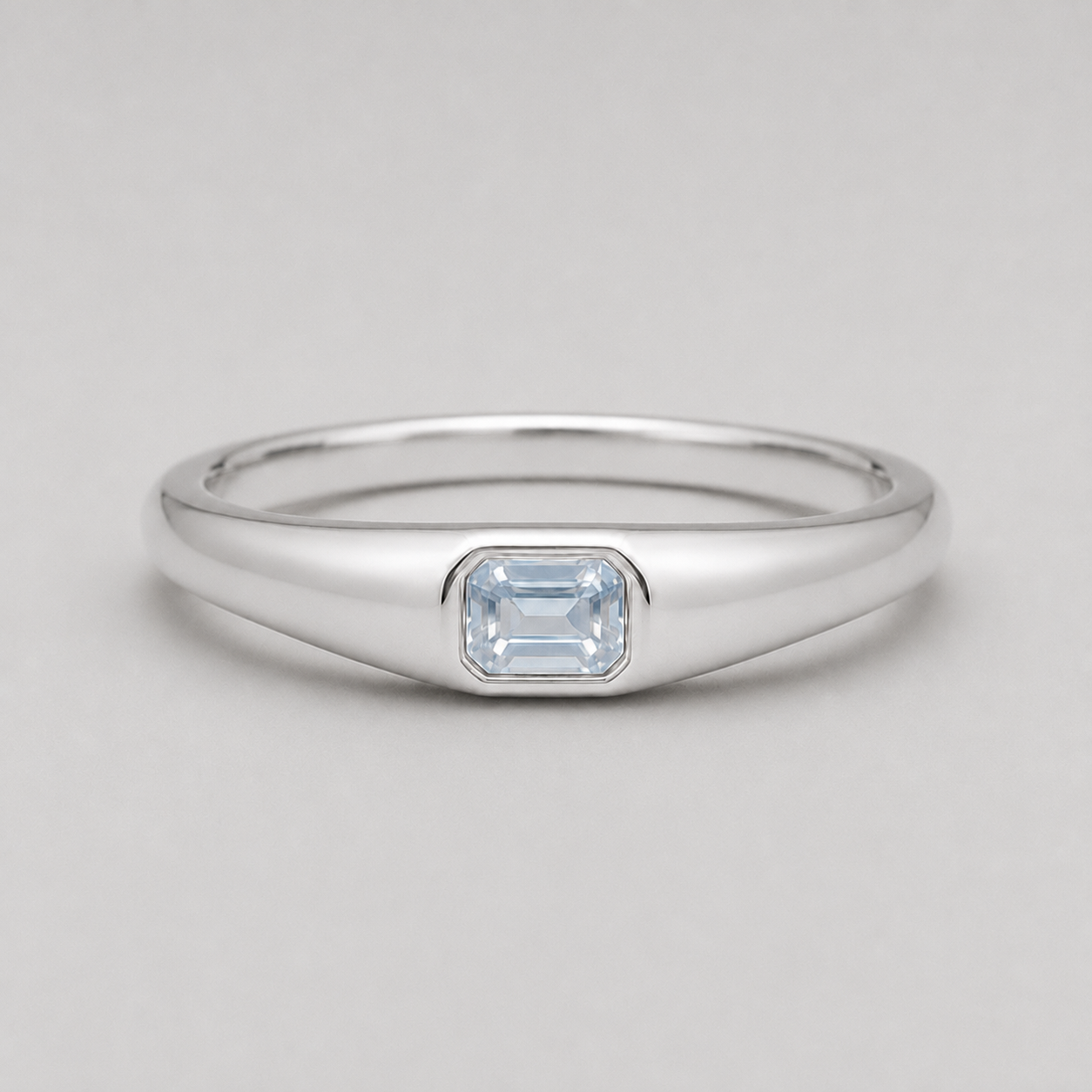 3D render of a polished silver ring featuring a rectangular emerald-cut white sapphire in a bezel setting, shown on a neutral light grey background.