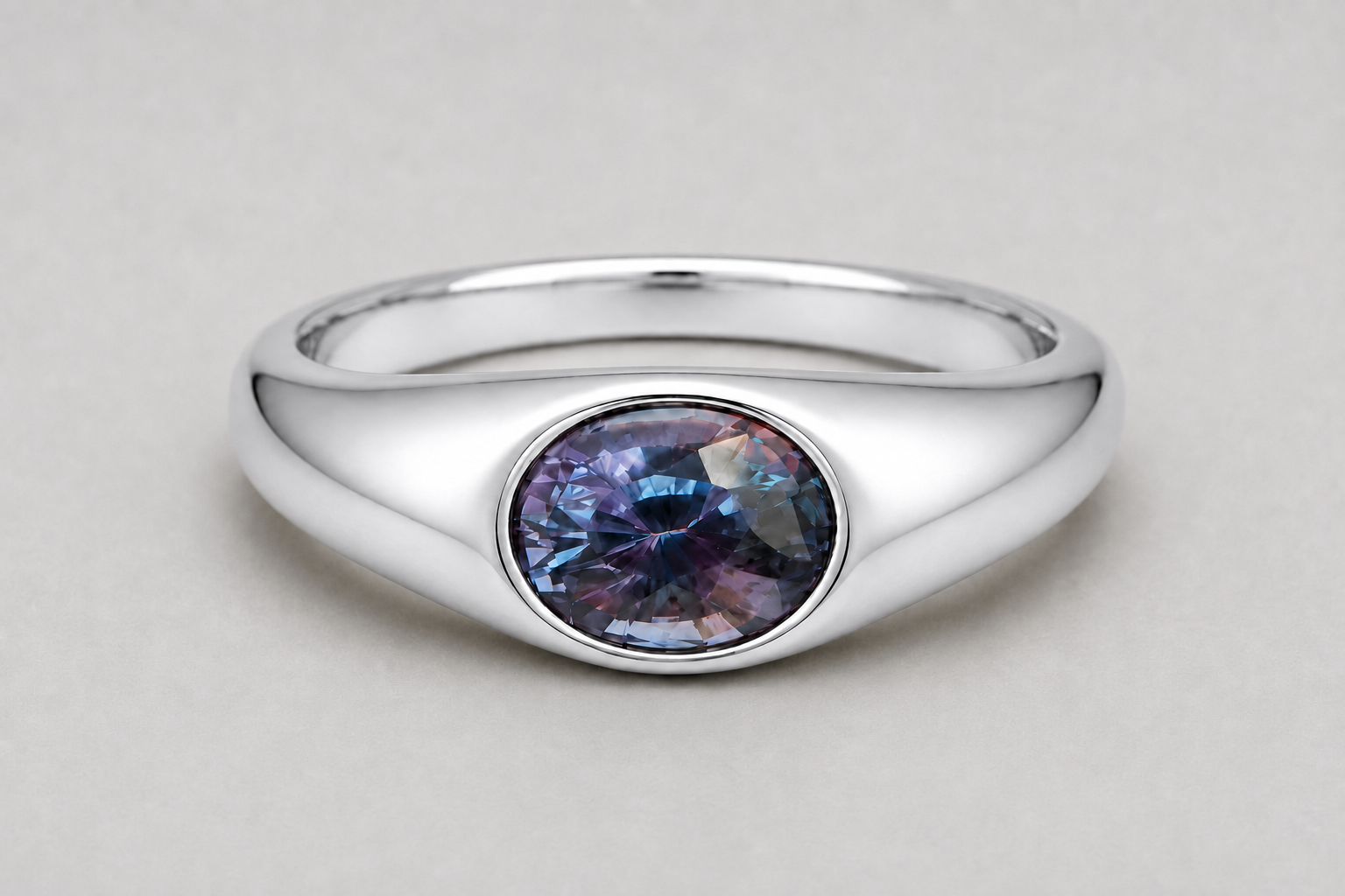 A 3D render of an oval-cut colour change sapphire flush set signet ring in a silver, shown on a neutral light grey background.