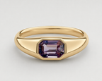 A yellow gold signet ring featuring a bezel-set emerald-cut purple spinel, shown on a neutral light grey background.