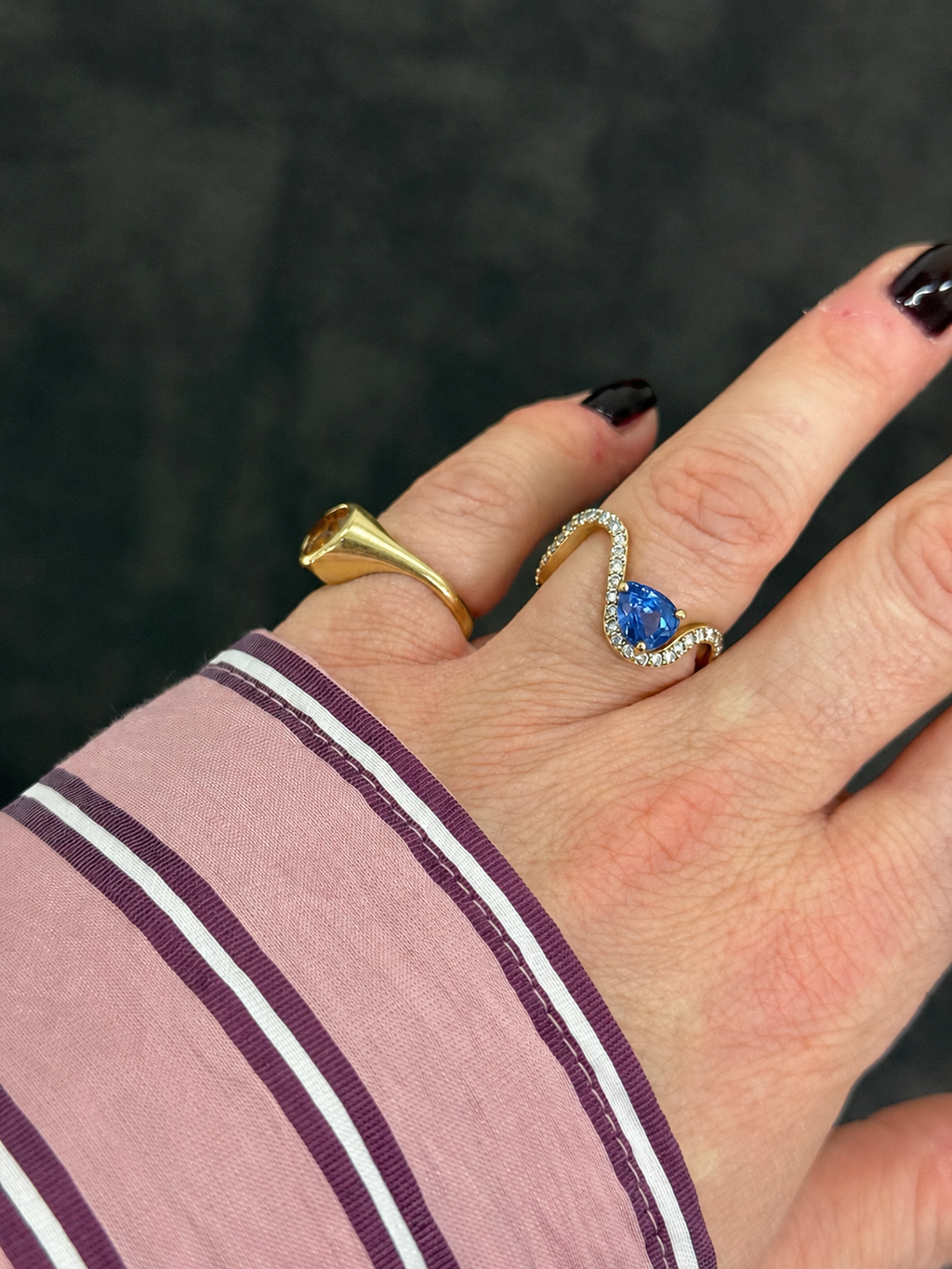 A close-up of a person's hand wearing two rings: a gold signet ring on the pinkie finger and a gold wave ring with a blue teardrop sapphire on the ring finger. The person is wearing a pink and purple striped long-sleeve shirt against a dark textured background.