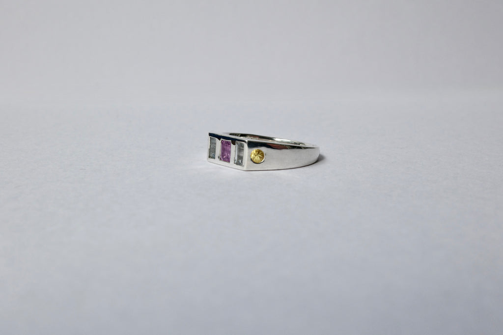 A silver ring with a rectangular bezel setting featuring a central purple stone, flanked by pale blue stones, and a small round yellow gemstone on the right, set against a plain light gray background.