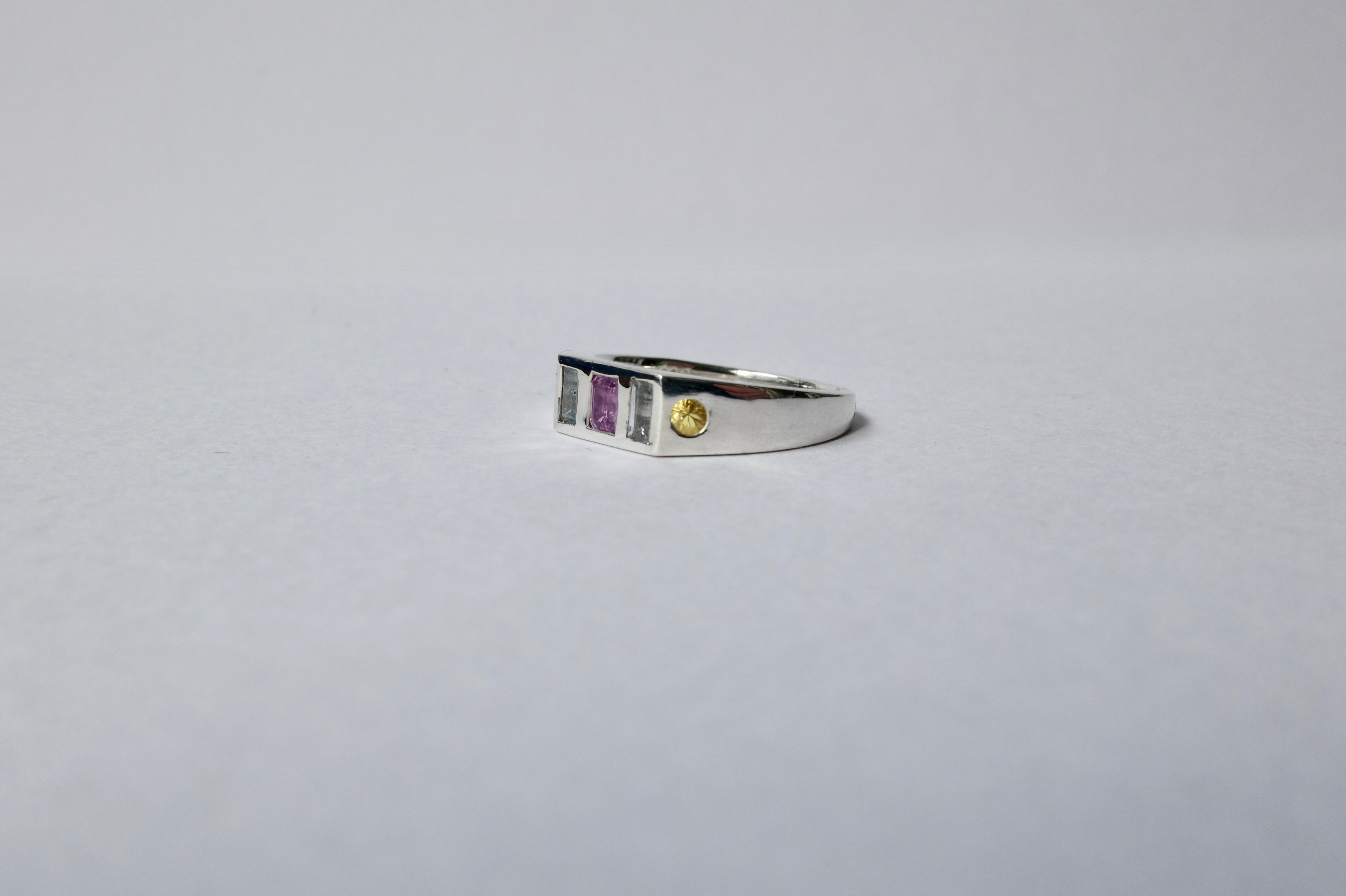 A silver ring with a rectangular bezel setting featuring a central purple stone, flanked by pale blue stones, and a small round yellow gemstone on the right, set against a plain light gray background.