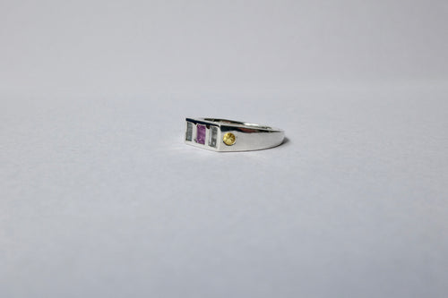 A silver ring with a rectangular bezel setting featuring a central purple stone, flanked by pale blue stones, and a small round yellow gemstone on the right, set against a plain light gray background.