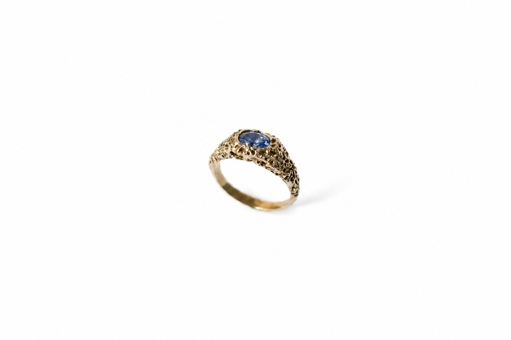 A textured gold ring featuring an openwork band and a central oval blue sapphire, shown on a plain white background.