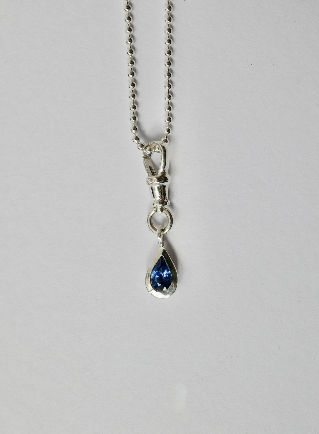 A silver teardrop pendant necklace with a deep blue sapphire, suspended from a silver ball chain against a light grey background.