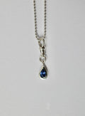 A silver teardrop pendant necklace with a deep blue sapphire, suspended from a silver ball chain against a light grey background.