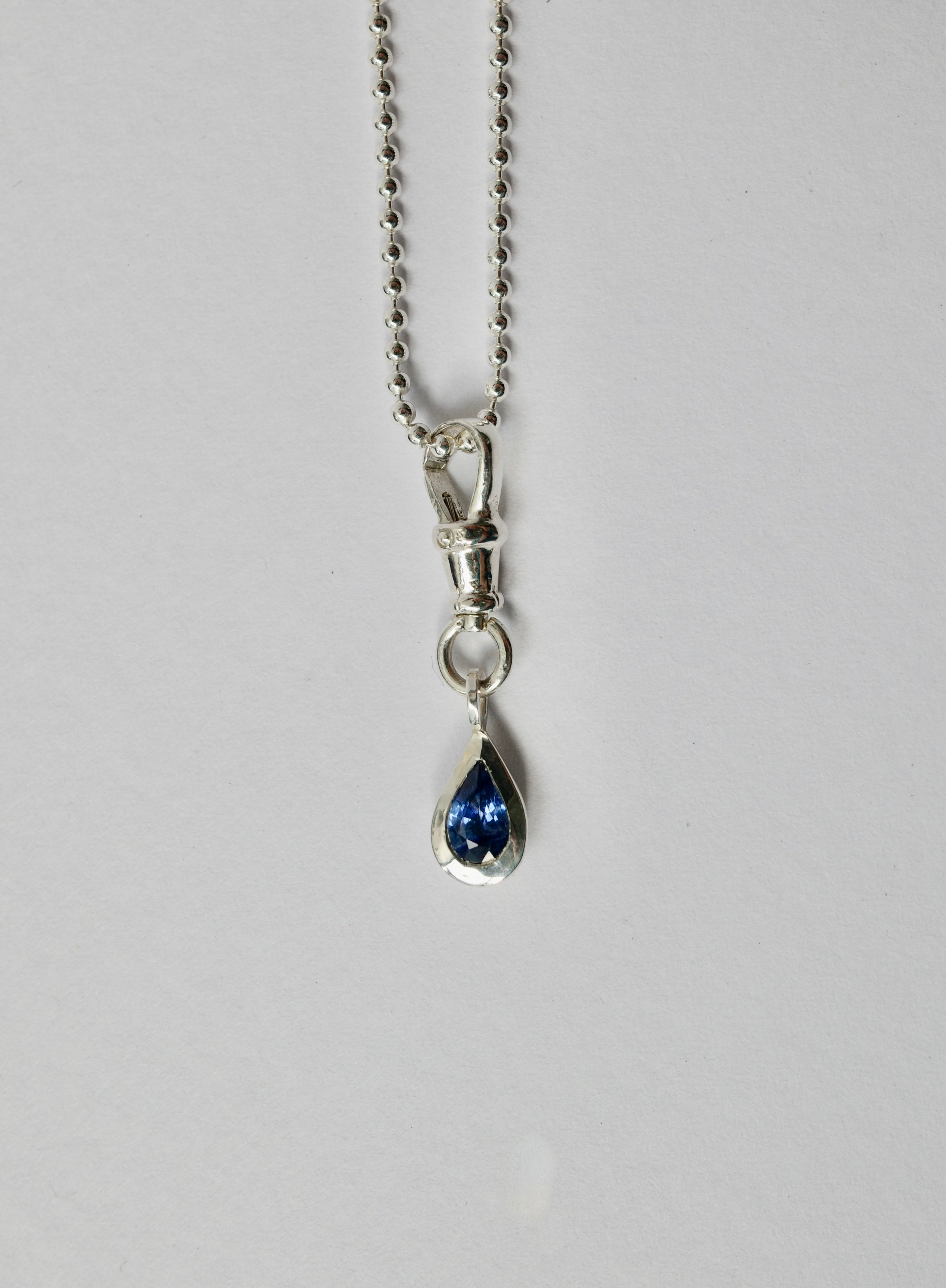 A silver teardrop pendant necklace with a deep blue sapphire, suspended from a silver ball chain against a light grey background.