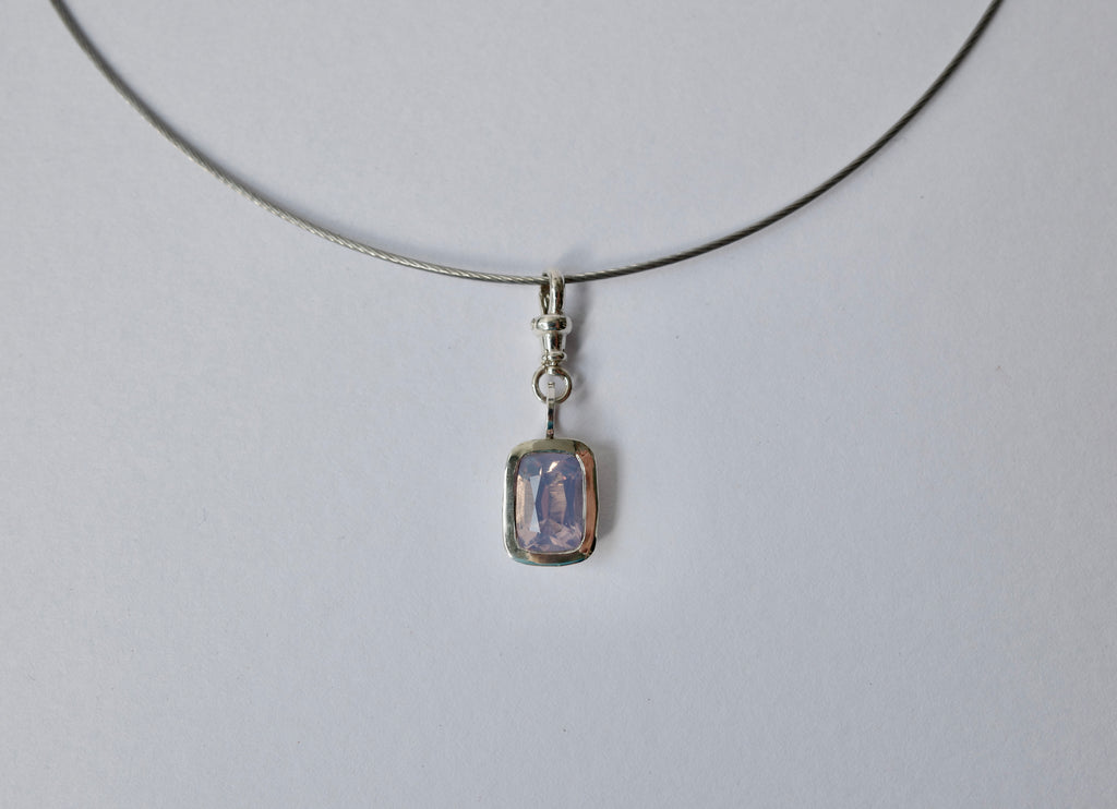 A delicate silver-toned necklace with a small rectangular pendant featuring a lavender-purple amethyst, shown against a plain light grey background.