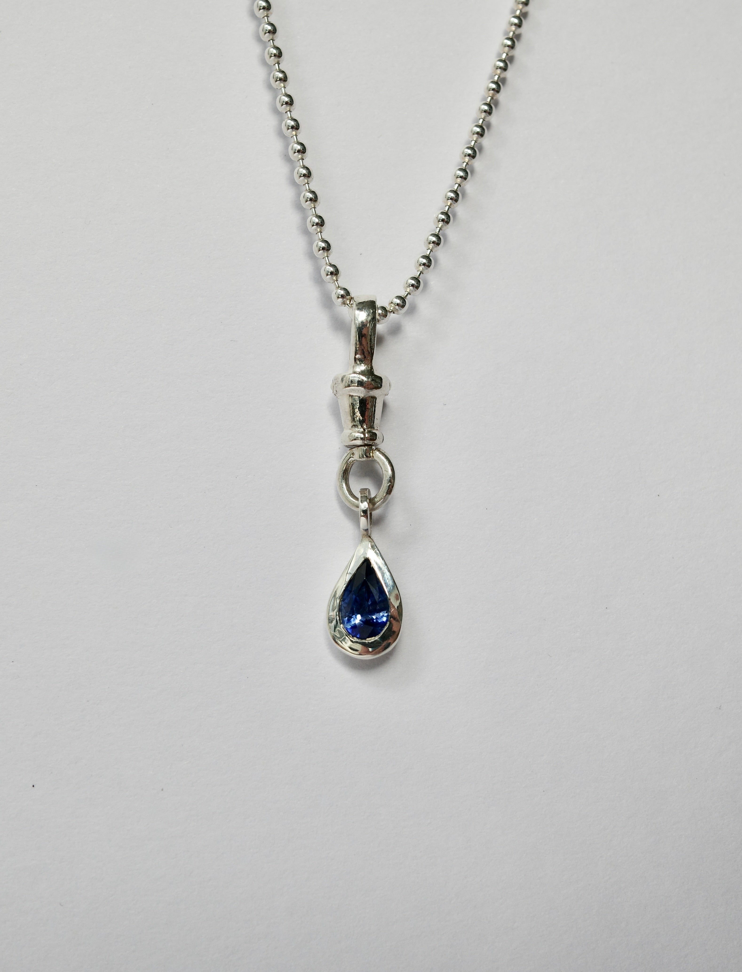 A silver ball chain necklace with a teardrop-shaped blue sapphire pendant, shown against a plain light grey background.