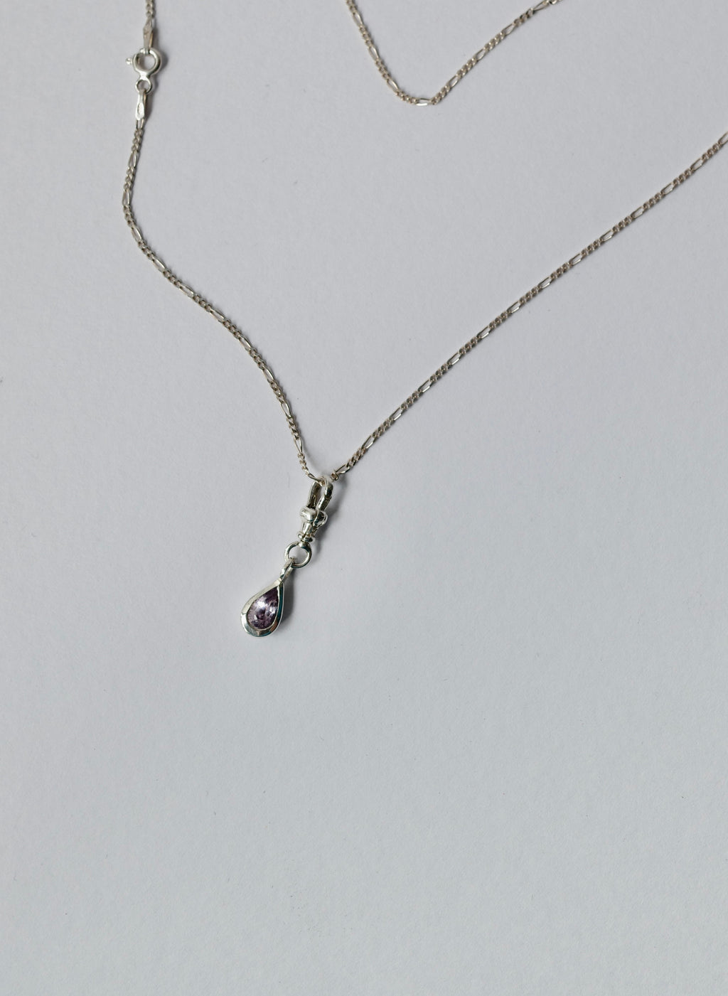 A silver chain necklace with a small teardrop-shaped pendant featuring a light purple sapphire, displayed on a light grey background.
