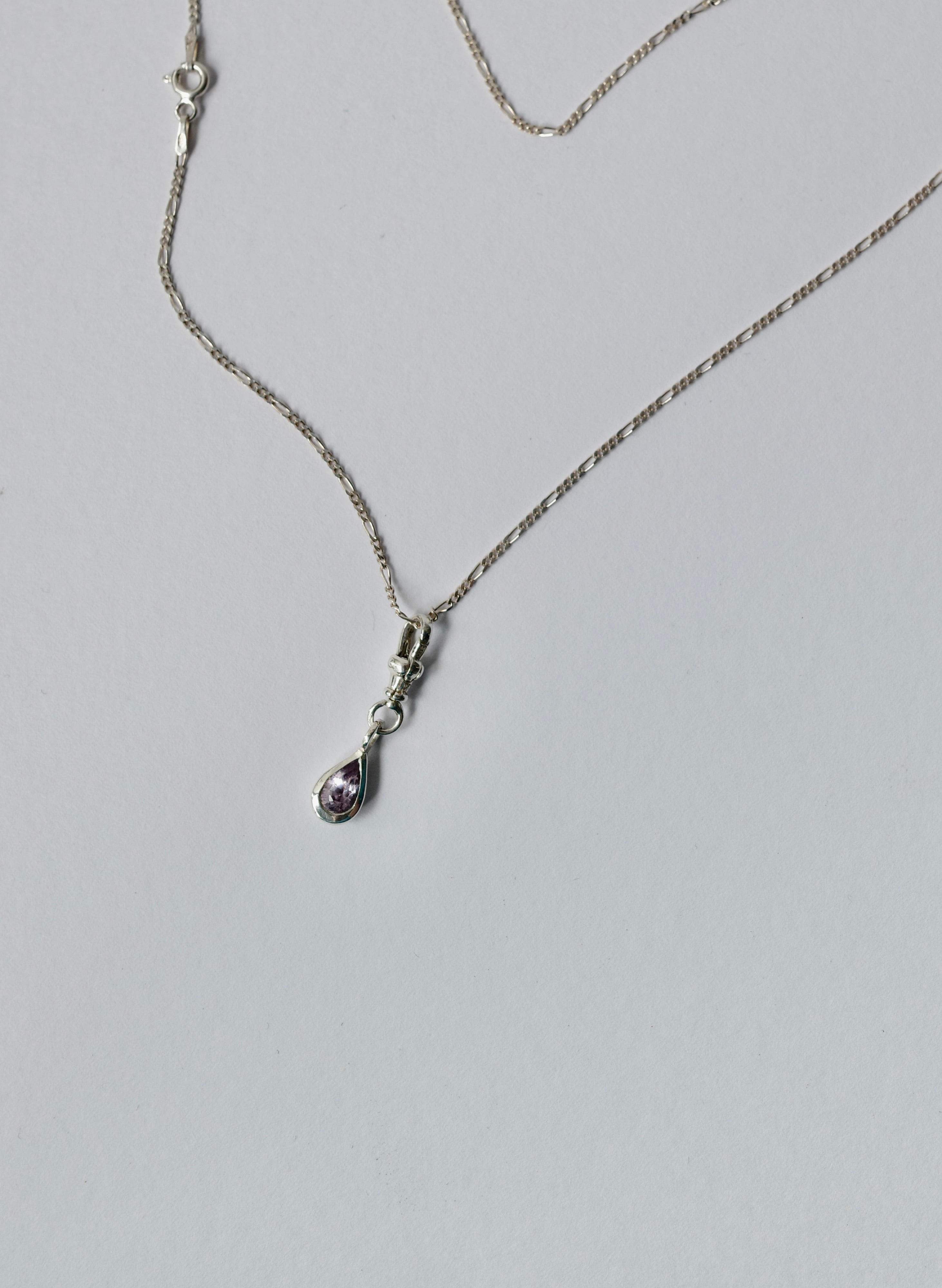 A silver chain necklace with a small teardrop-shaped pendant featuring a light purple sapphire, displayed on a light grey background.