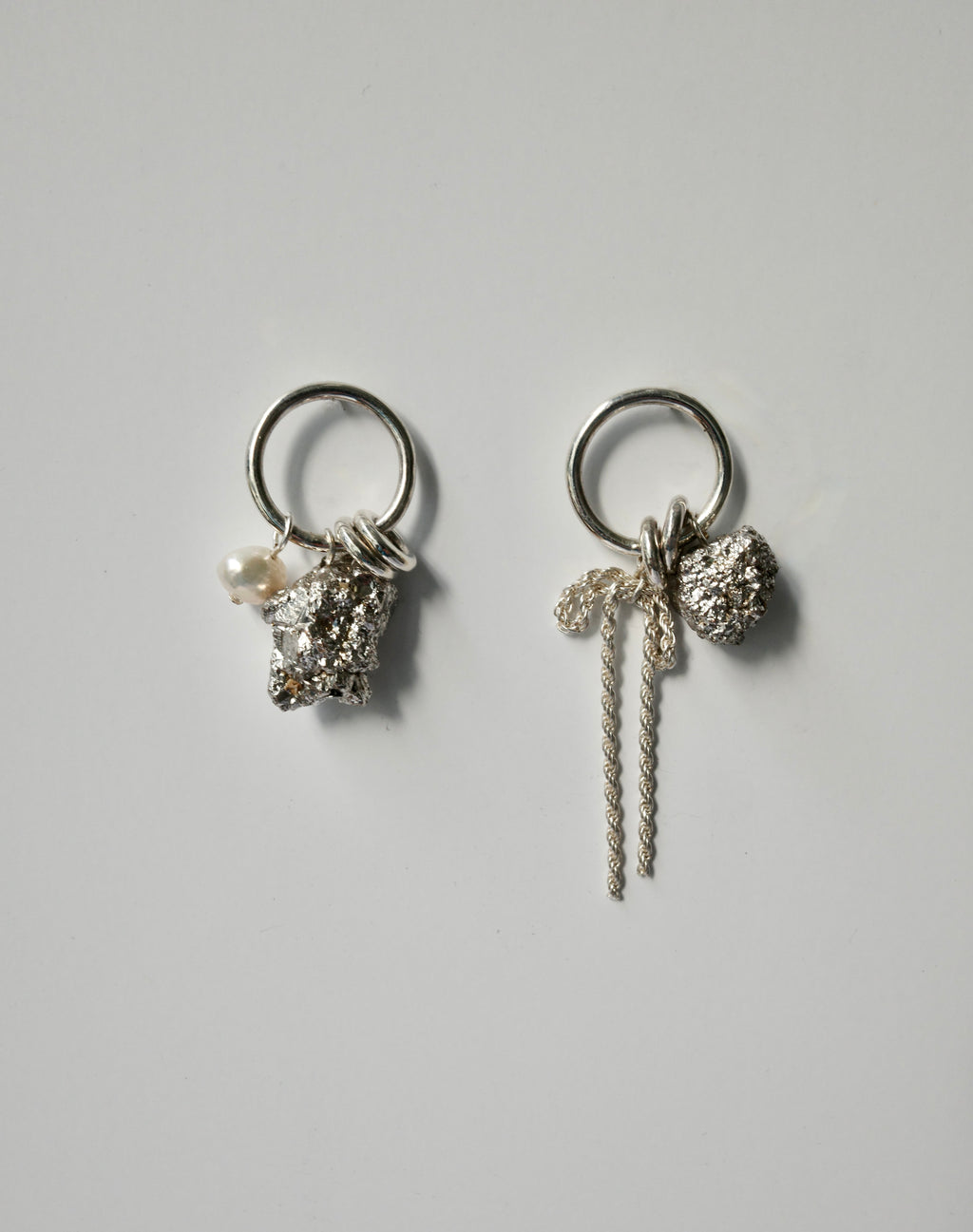 A pair of silver-toned dangle earrings on a light gray background. Each earring features a circular ring, a textured silver charm, a small white pearl, and a long twisted silver rope detail.