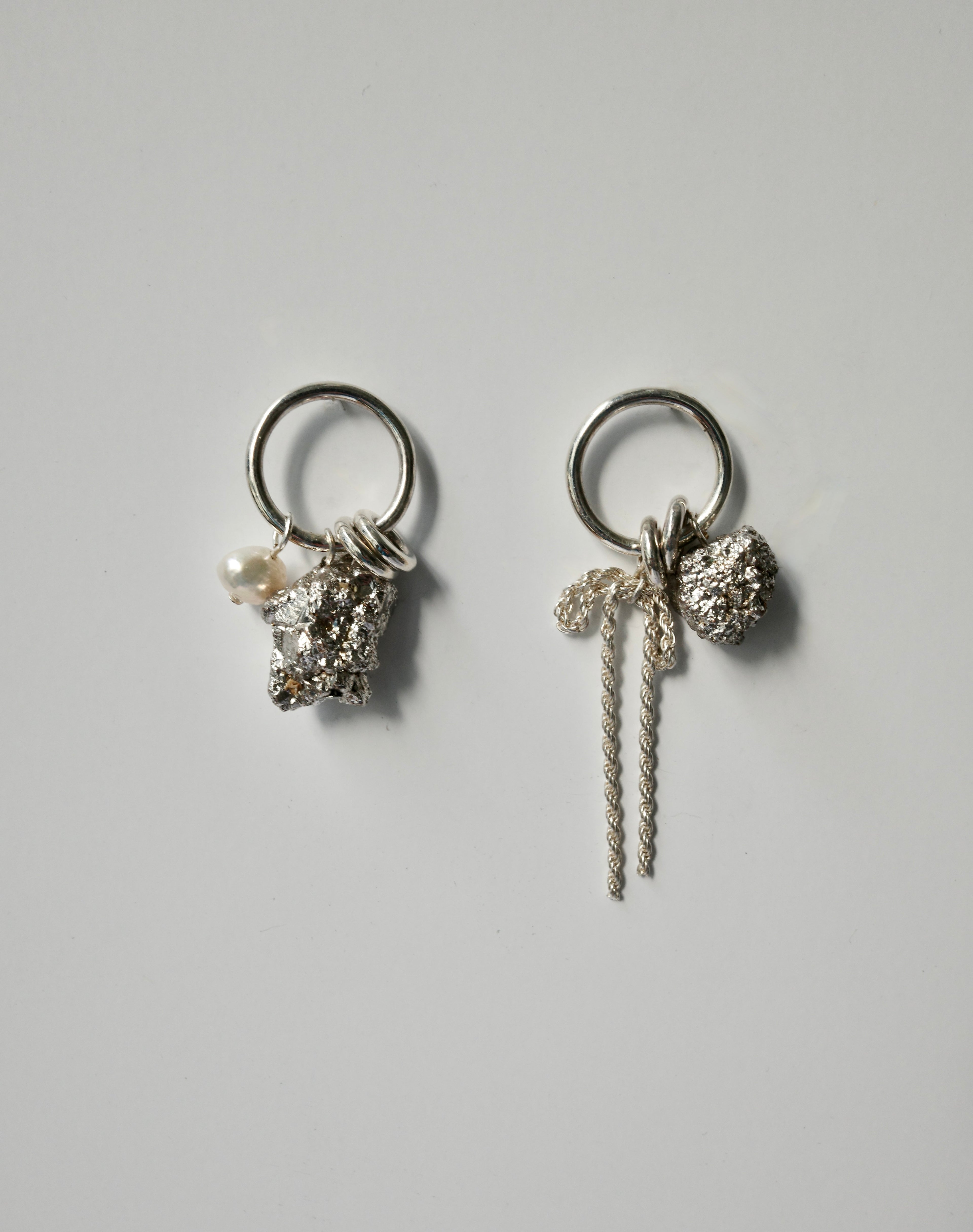 A pair of silver-toned dangle earrings on a light gray background. Each earring features a circular ring, a textured silver charm, a small white pearl, and a long twisted silver rope detail.