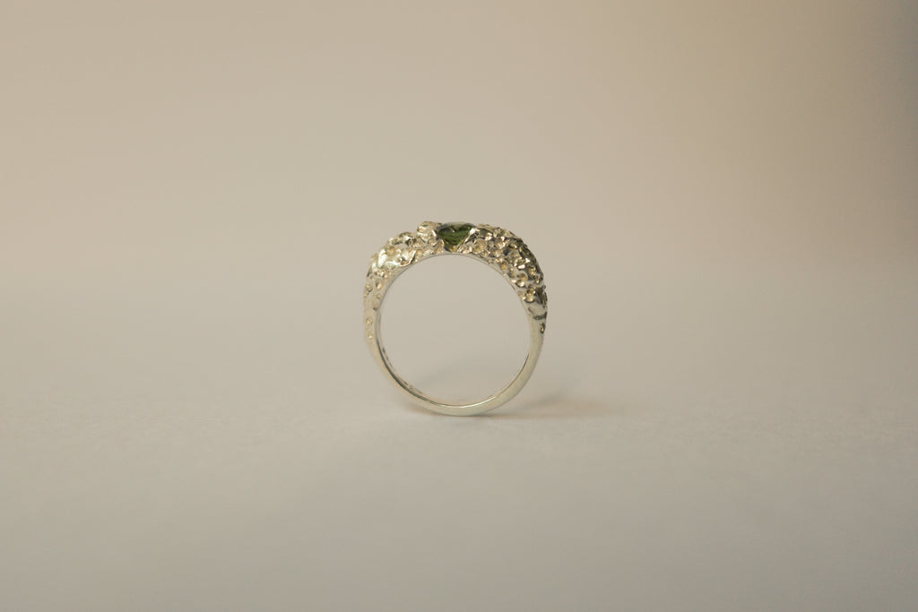 A silver textured ring with a small green sapphire, photographed against a plain light beige background.