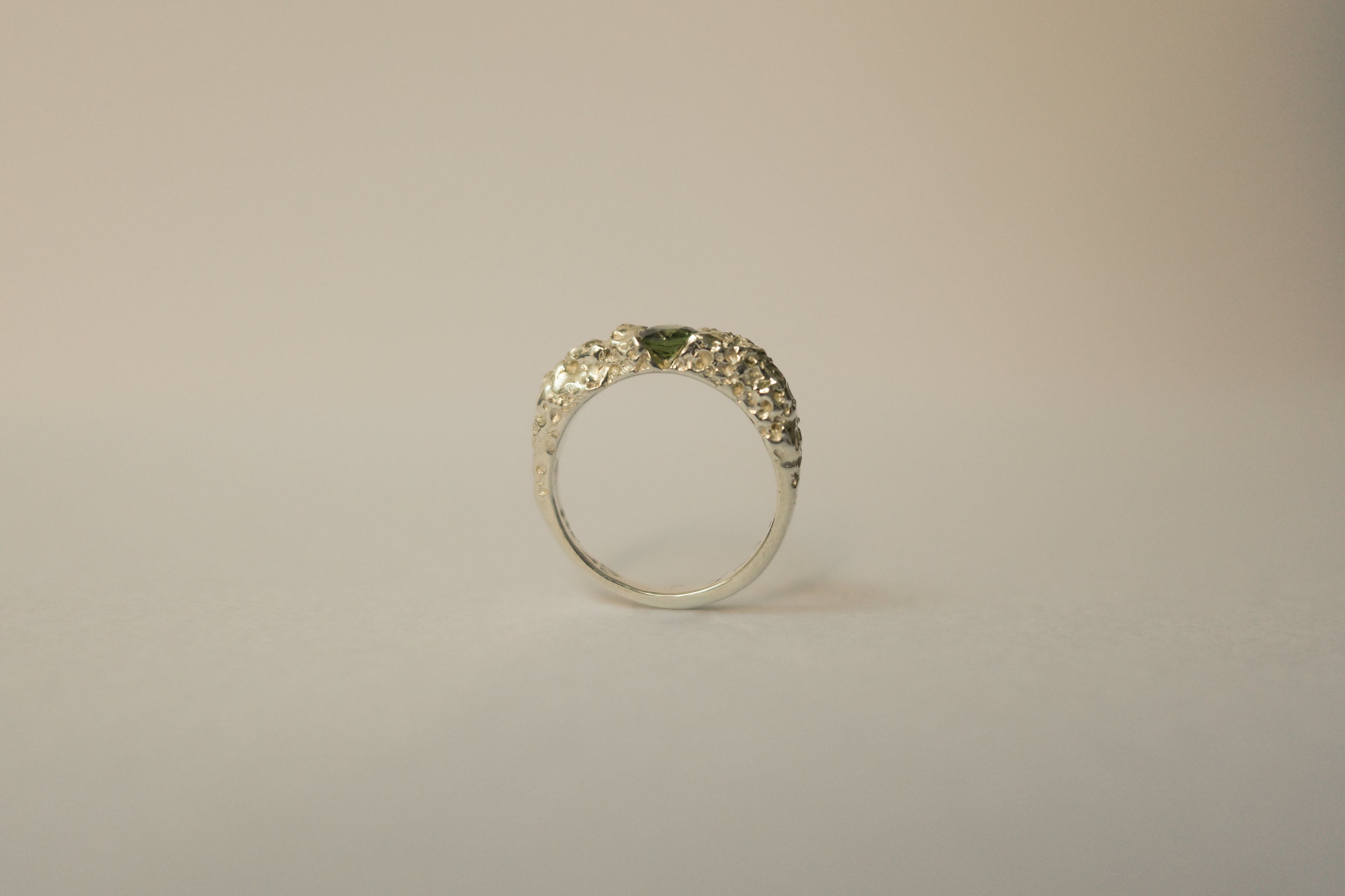 A silver textured ring with a small green sapphire, photographed against a plain light beige background.