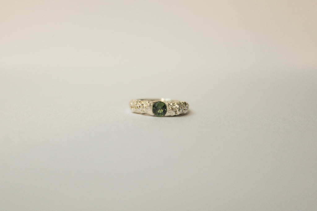 A textured silver ring featuring a central round green sapphire flanked, set against a plain light grey background.