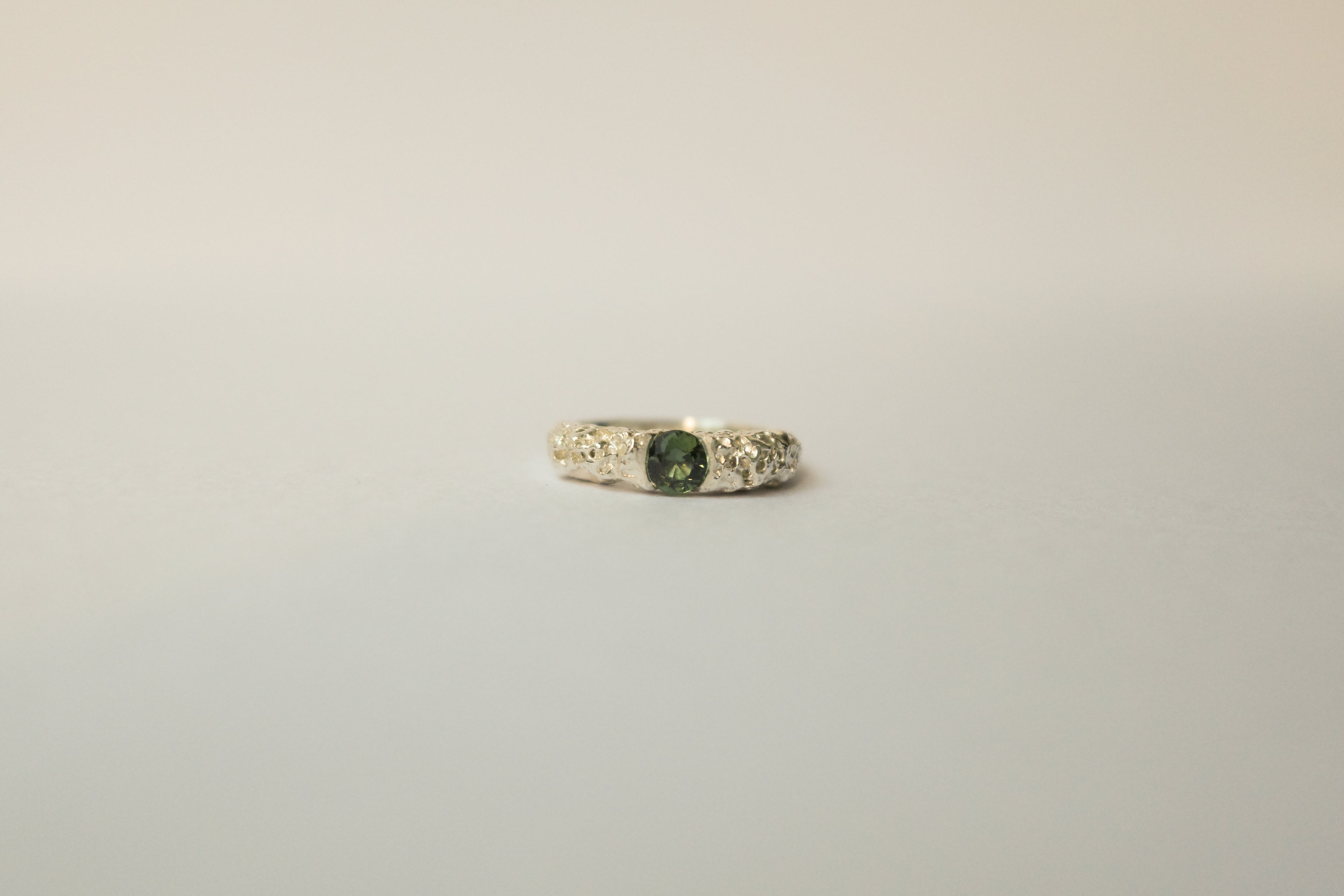 A textured silver ring featuring a central round green sapphire flanked, set against a plain light grey background.