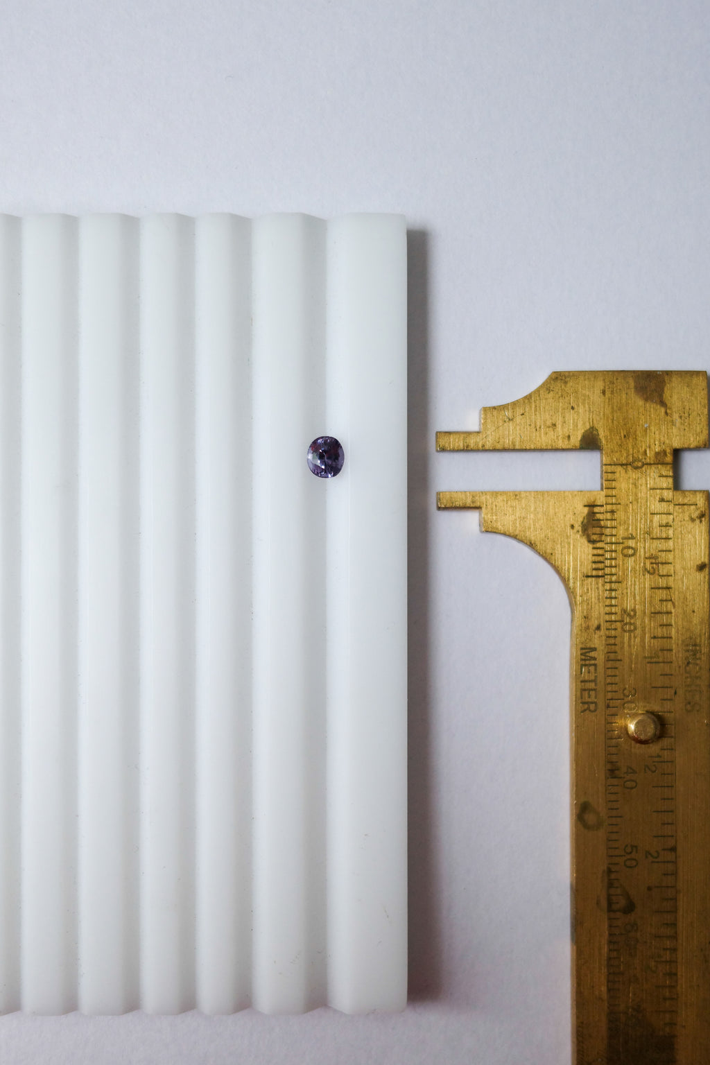 A close-up of an oval colour change sapphire set in a gemstone tray, shown next to a brass calliper against a plain light background.