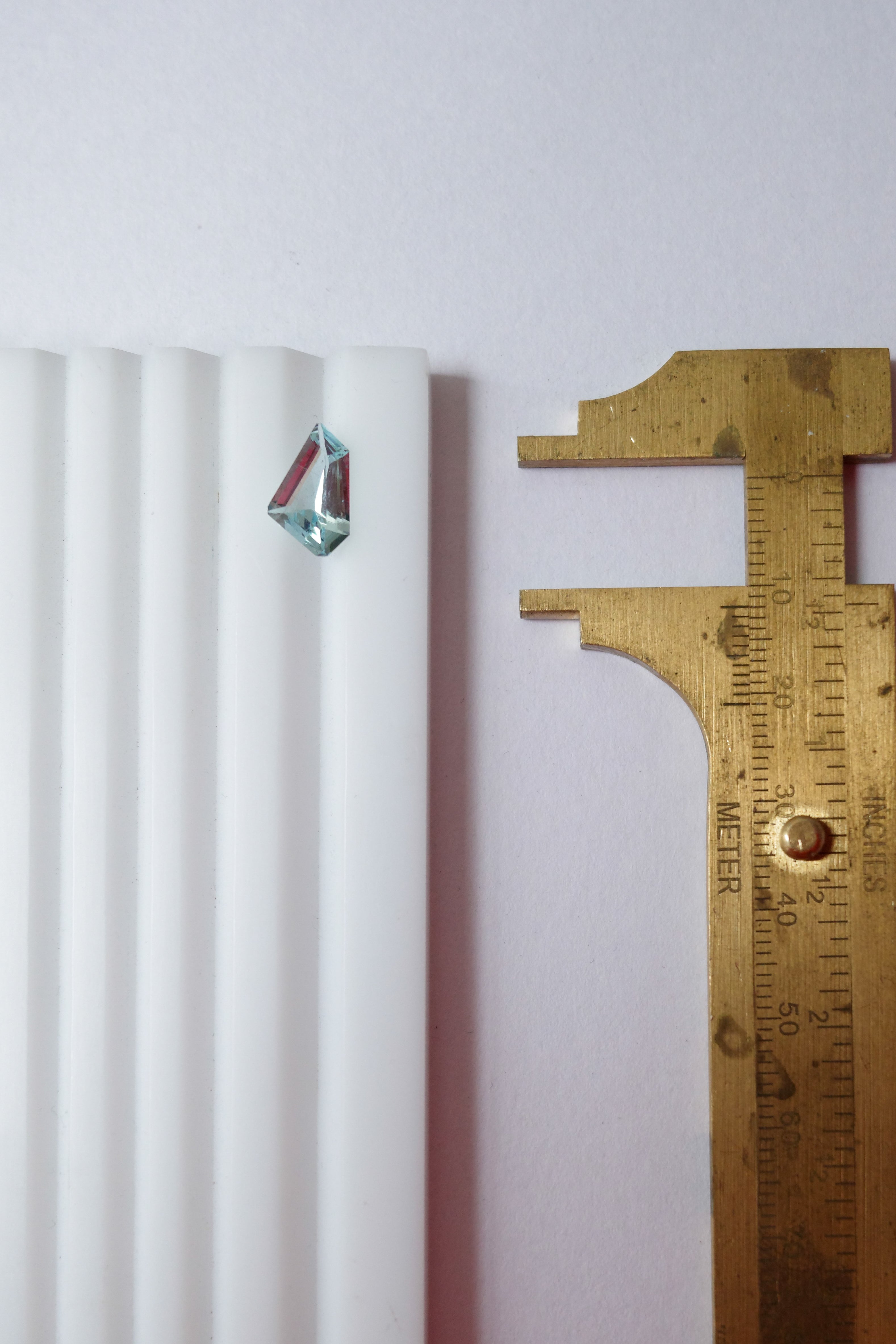 A loose freeform light blue aquamarine on a gemstone sorting tray next to brass callipers for size and scale context