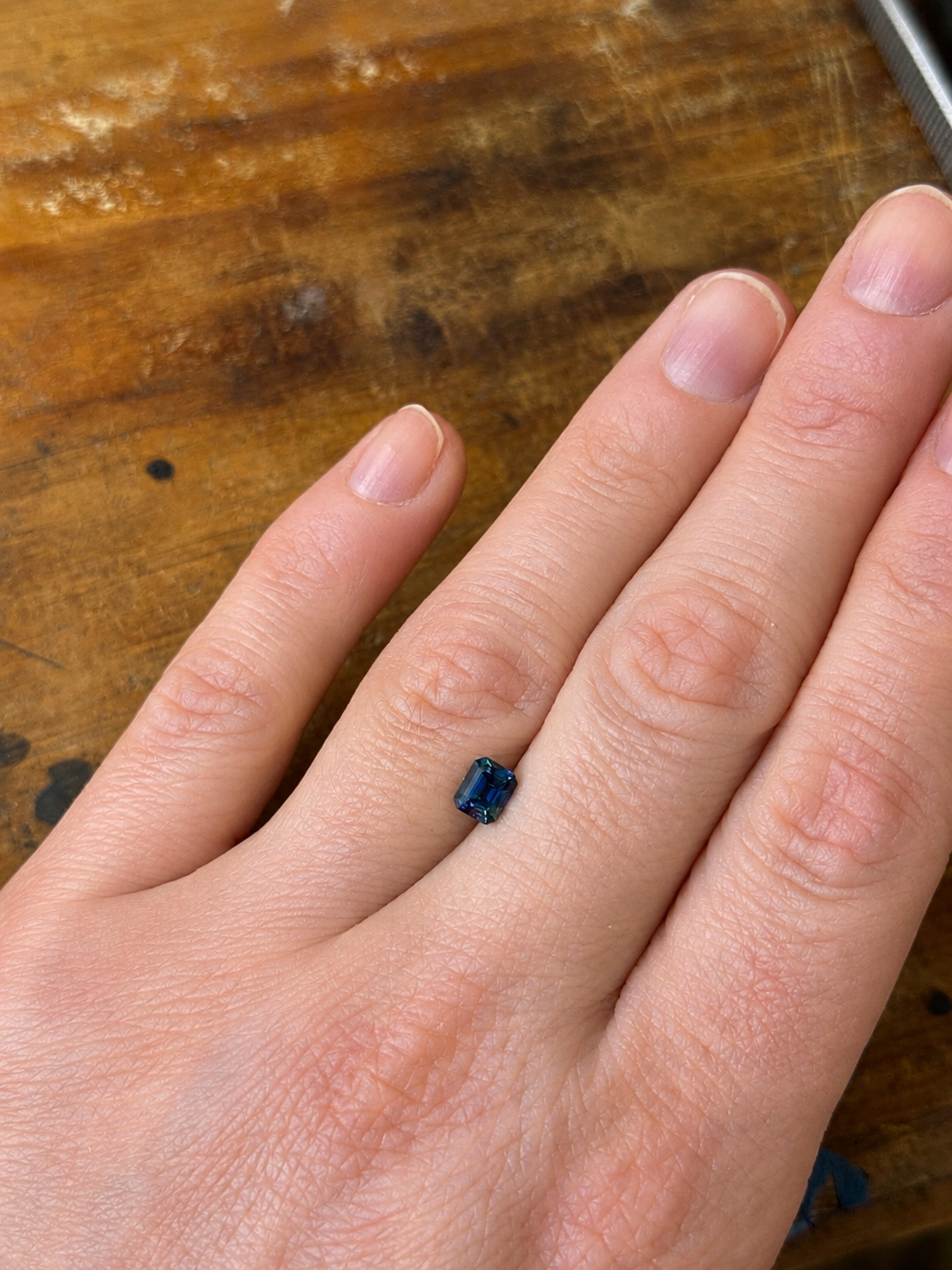 A loose blue/green emerald cut parti sapphire on a persons hand for scale
