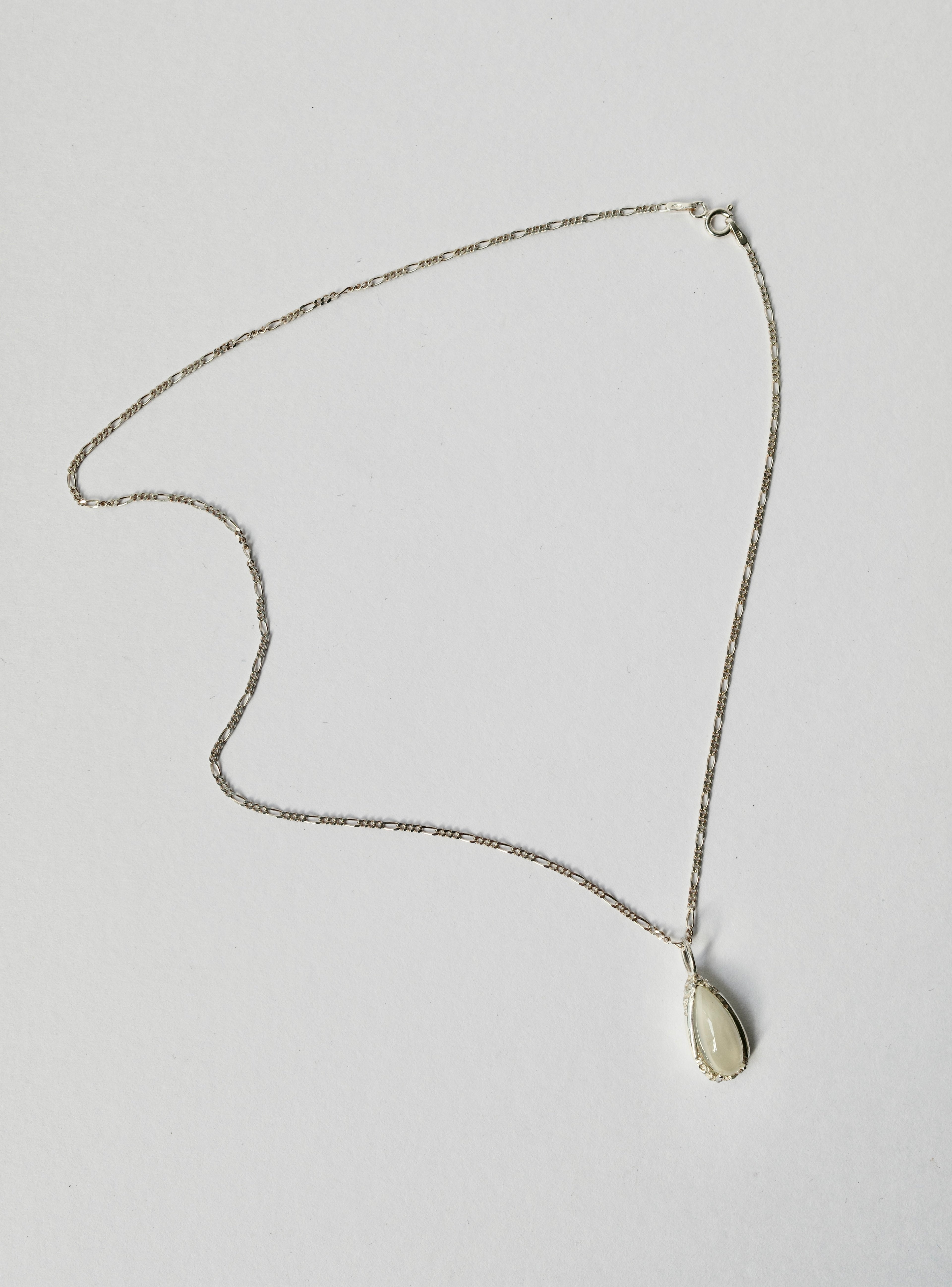 A delicate silver chain necklace with a small, teardrop-shaped milky white moonstone pendant, displayed on a plain light grey background.