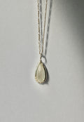 A silver necklace with a teardrop-shaped pendant featuring a milky white moonstone, hanging against a neutral grey background.
