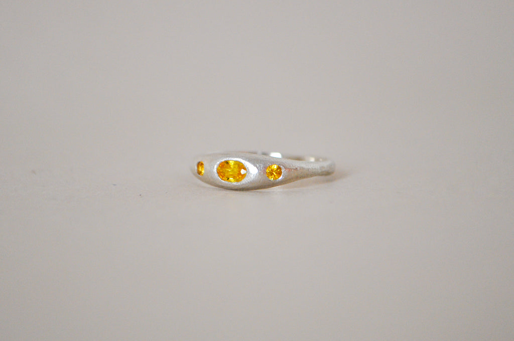 A silver ring featuring a central oval yellow sapphire flanked by two smaller round yellow sapphire, set against a plain light grey background.