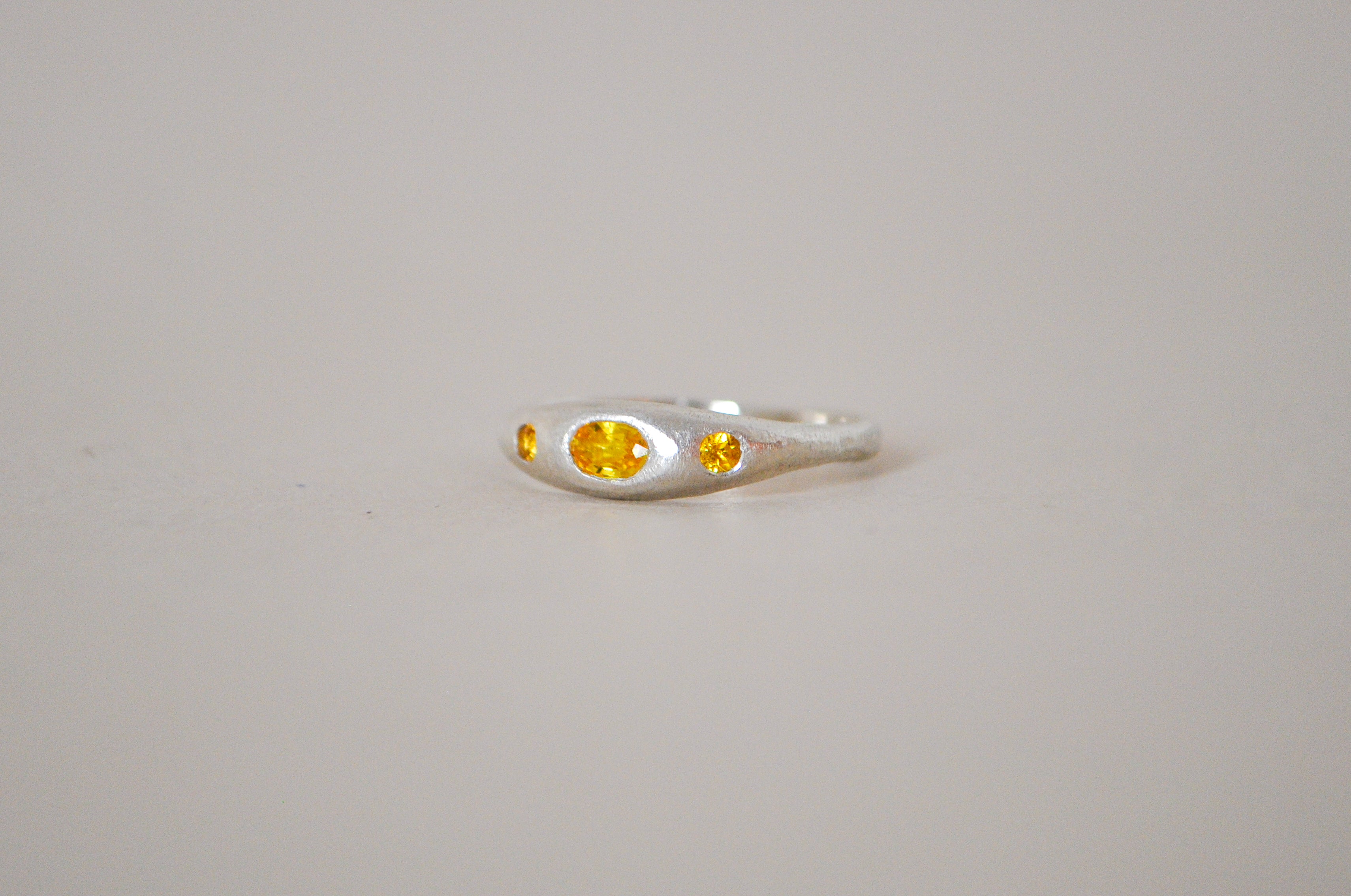 A silver ring featuring a central oval yellow sapphire flanked by two smaller round yellow sapphire, set against a plain light grey background.