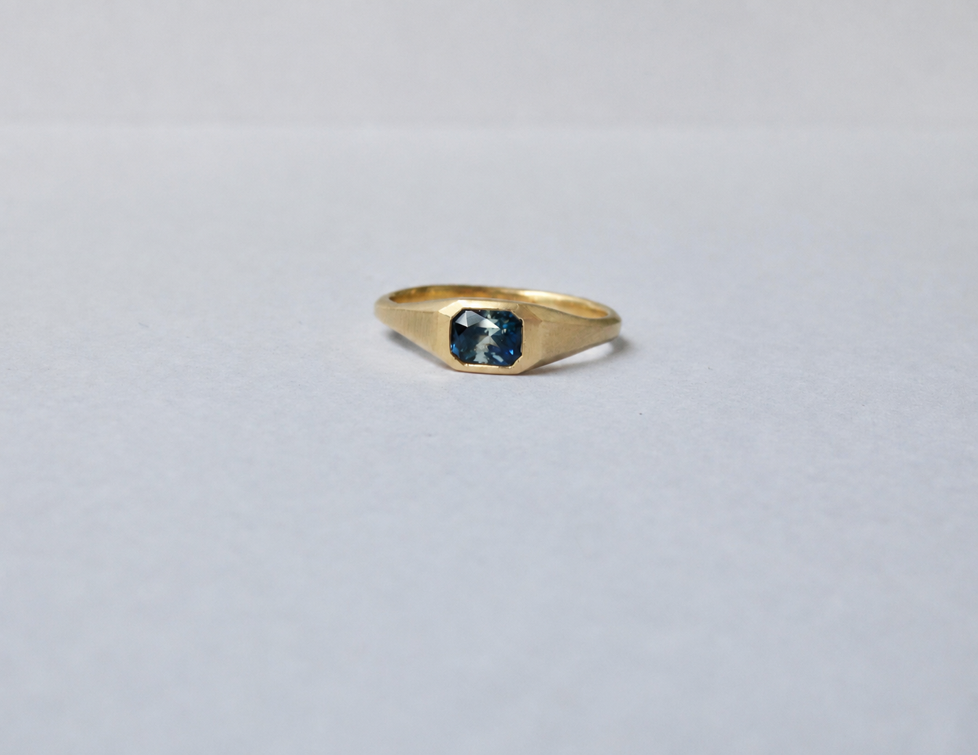 A gold signet ring with a square-cut parti sapphire, displayed on a plain light grey background.