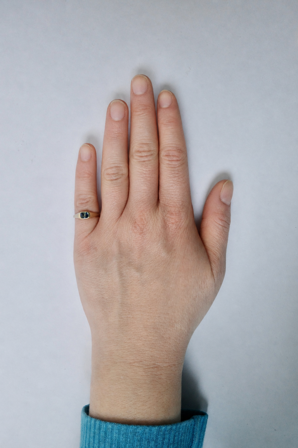 A close-up of a person's hand wearing a gold pinkie ring with a small parti sapphire, set against a plain light grey background.