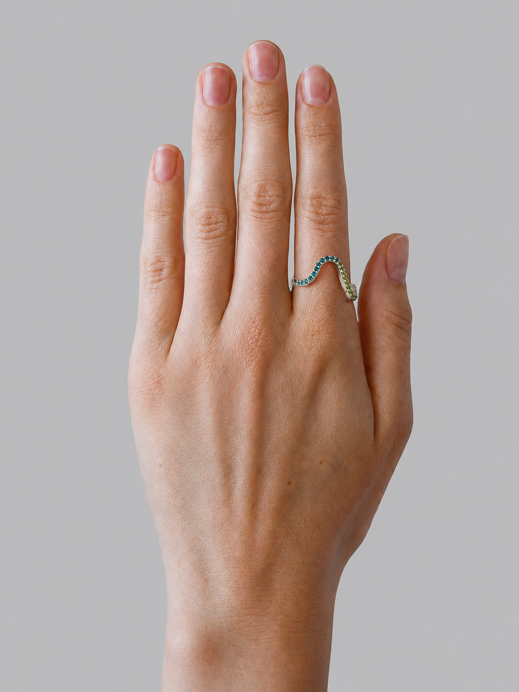 A close-up of a hand wearing a wavy ring with small blue and green gemstones on a plain grey background.