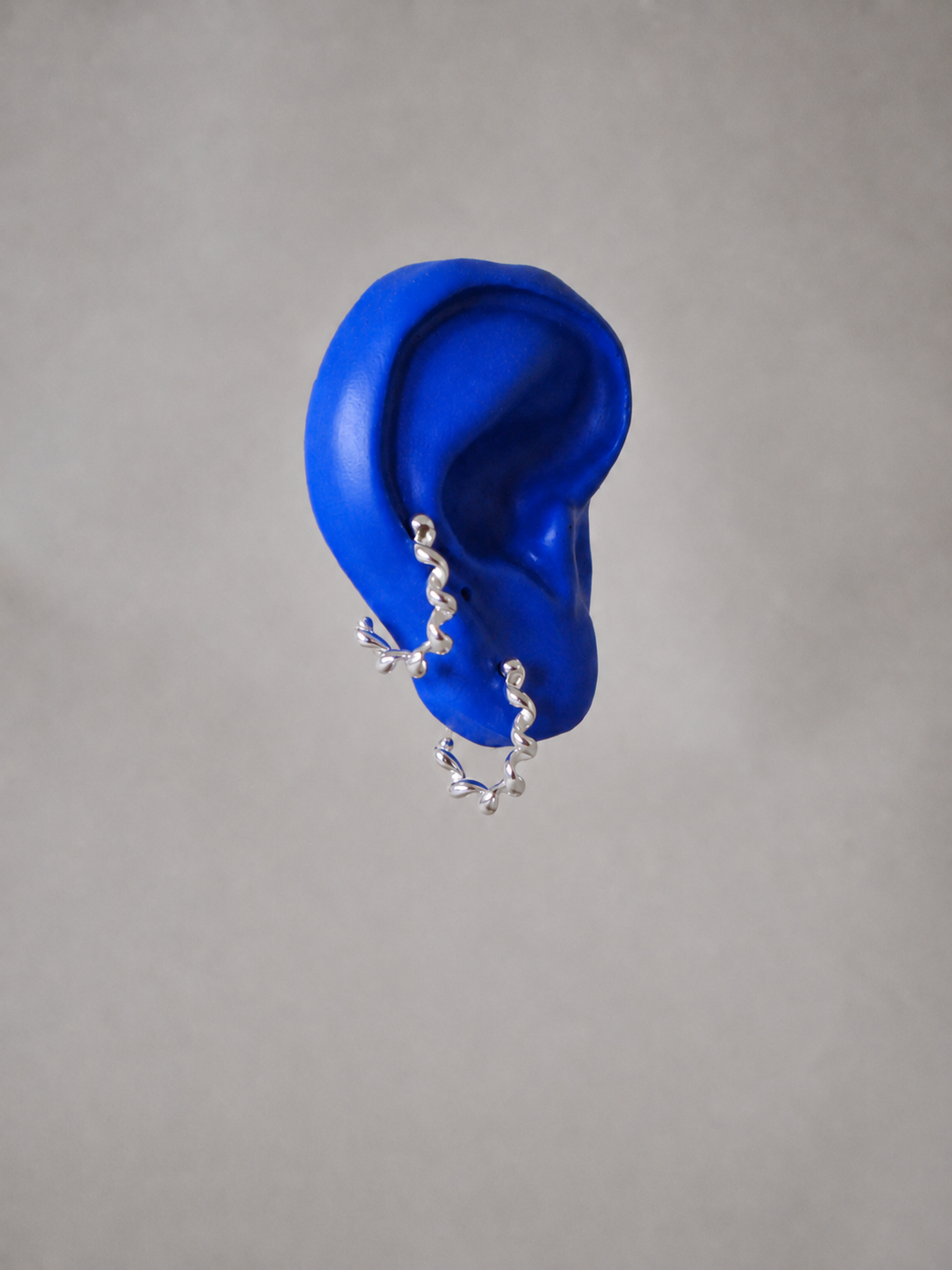 A pair of silver spiral hoop earrings, displayed on a bright blue ear against a neutral grey background.