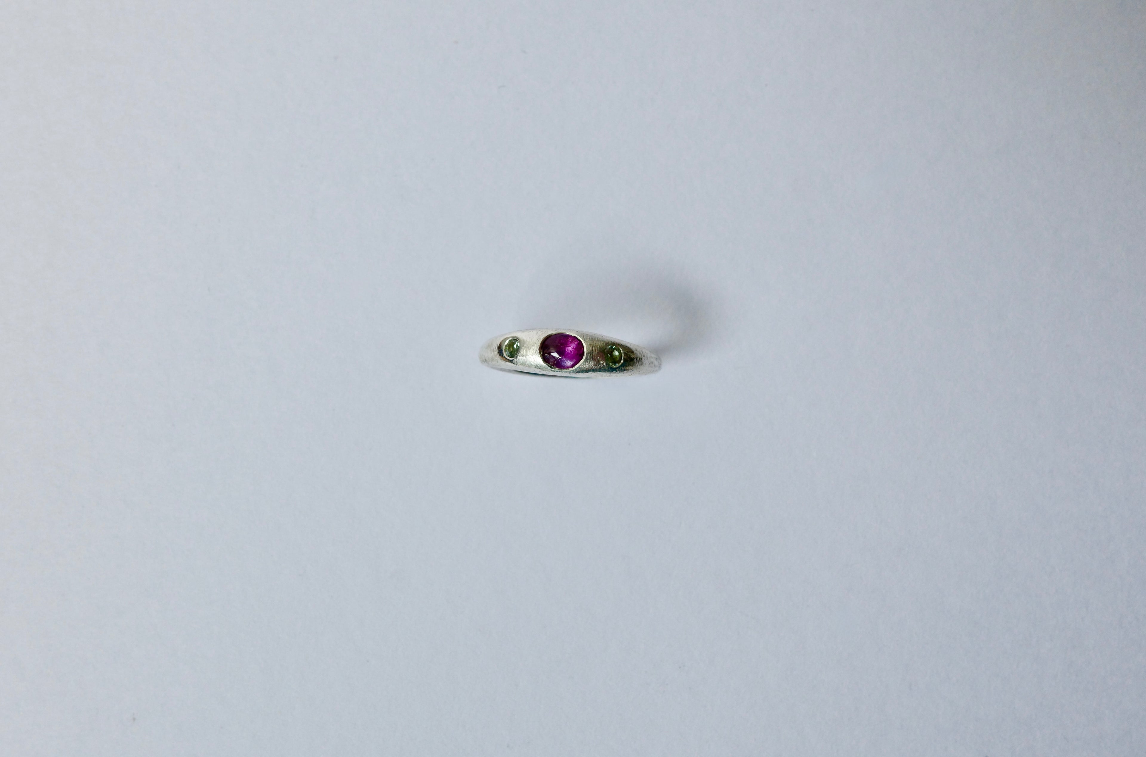 A delicate silver ring featuring a central round purple cabochon sapphire flanked by two tiny green sapphires, set against a plain light grey background.