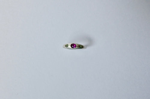 A delicate silver ring featuring a central round purple cabochon sapphire flanked by two tiny green sapphires, set against a plain light grey background.
