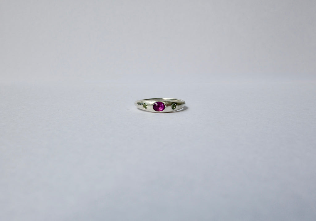 A delicate sterling silver ring featuring a central oval purple cabochon sapphire accented by tiny green sapphires on either side, set against a plain light grey background.