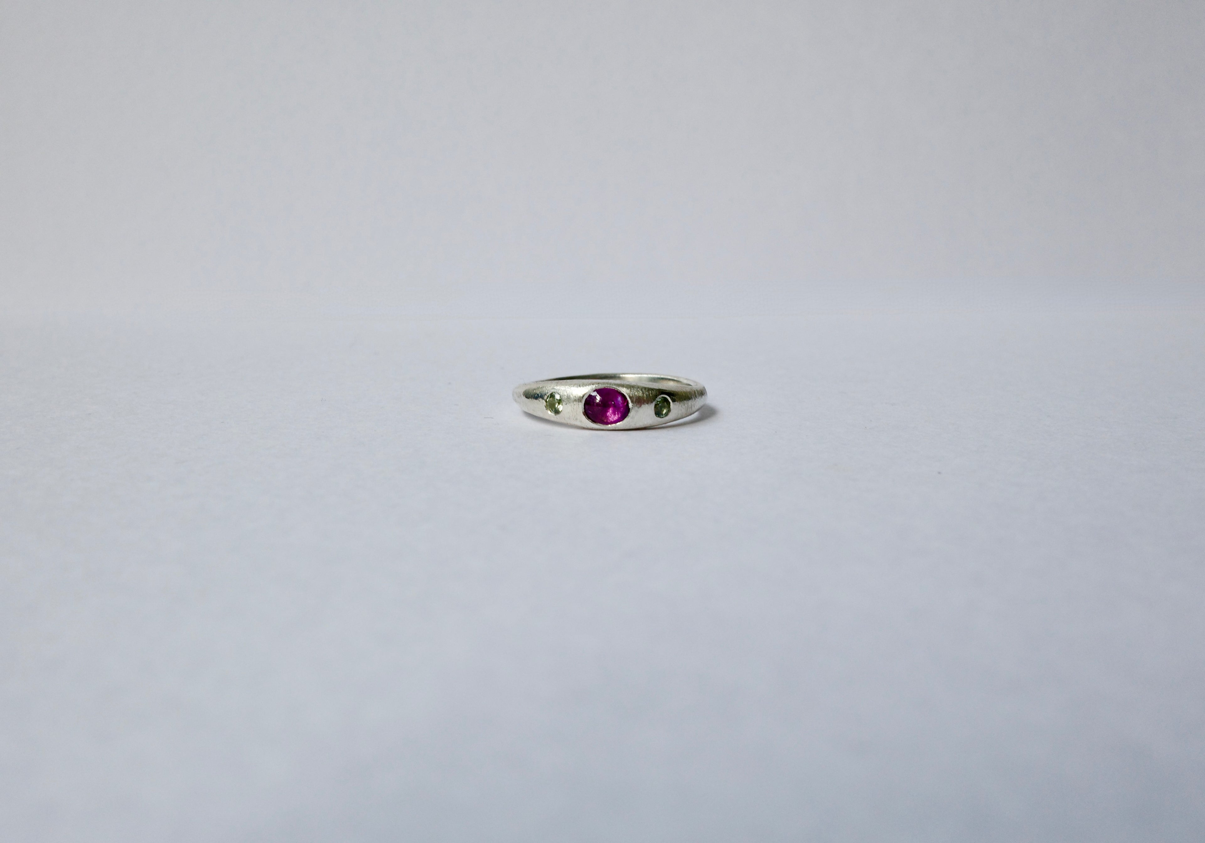 A delicate sterling silver ring featuring a central oval purple cabochon sapphire accented by tiny green sapphires on either side, set against a plain light grey background.