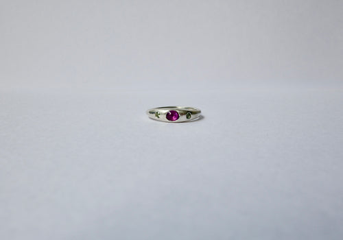 A delicate sterling silver ring featuring a central oval purple cabochon sapphire accented by tiny green sapphires on either side, set against a plain light grey background.